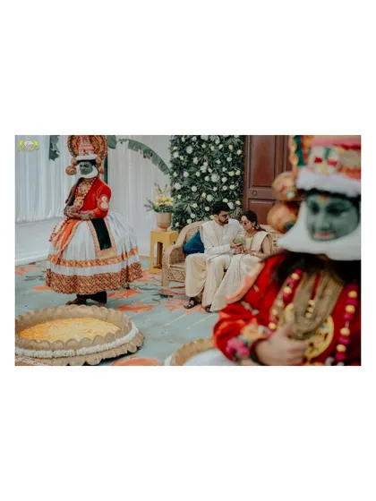 An artistic composition with the Kathakali dancer in the foreground. This shot creates depth and tells a story, capturing a quiet moment between the couple amidst the cultural festivities.