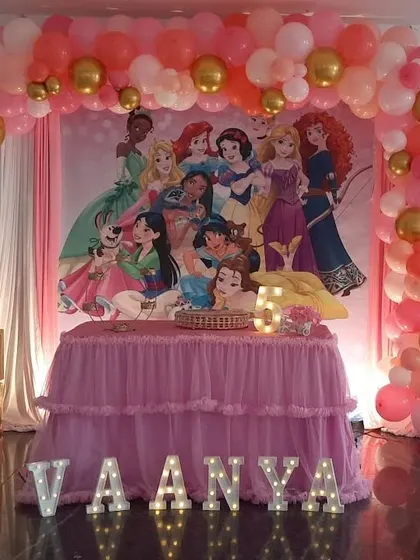 A Disney Princess theme featuring a large backdrop with multiple princesses. The decoration is completed with a pink and gold balloon arch and marquee letters.