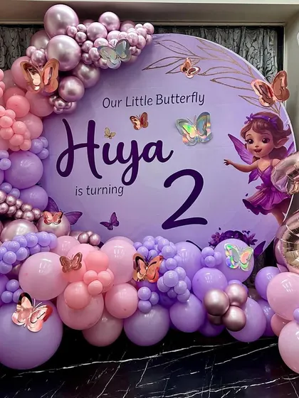 A magical butterfly-themed second birthday for "Hiya," featuring a large circular backdrop in soft purple. The decor is brought to life with a stunning organic balloon garland in shades of pink and purple, accented with metallic and iridescent butterfly cutouts and a large rose gold '2' foil balloon.