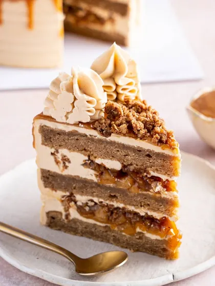 A slice of our Apple and Salted Caramel Cake, a true hug in cake form. You can see the layers of spiced apple sponge, gooey apple pie filling, and silky salted caramel buttercream, all topped with a crunchy crumble.