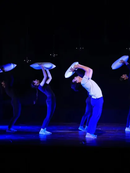 Dancers in "ME & MY PILLOW" perform a synchronized backbend while holding pillows. This moment of unity and physical expression is set against a dark stage with dramatic blue lighting.