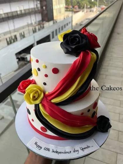 A vibrant two-tier cake celebrating Uganda's 60th Independence Day. The design incorporates the colors of the Ugandan flag in drapes and fondant roses, with polka dots adding a festive touch.