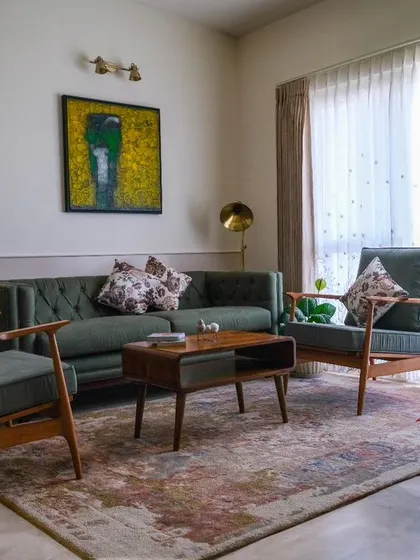 The living room of the 'Earthy Chic Bachelorette Pad,' where a Forest Green tufted sofa and traditional teak wood lounge chairs create a delightful fusion of warmth and earthy ambiance against a soft white paneled wall.