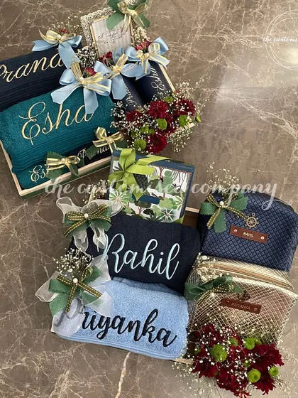 We create unique hampers that blend practicality with sentiment. This display shows two distinct couple sets, one in blue and green tones and another in blue and gold, each with personalized towels and matching accessories.