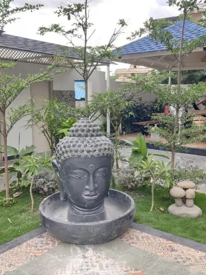 A large Buddha head sculpture converted into a water fountain, placed in the corner of a garden. The water flows gently from the top into the basin below.