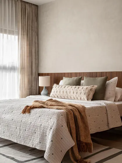 The master bedroom in the Warm Minimal home features lime-washed walls and a wood-paneled headboard. The layered neutral bedding and soft lighting create a deeply restful and elegant space.