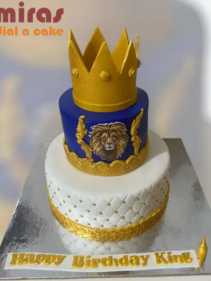 A regal two-tier cake for a "Birthday King." The design combines a quilted white bottom tier with a royal blue top tier featuring a lion crest and a golden crown.
