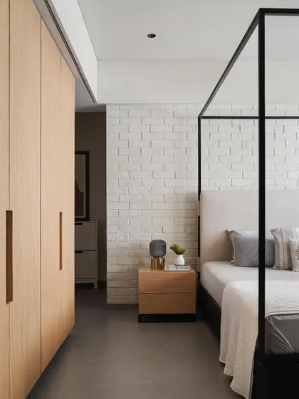 In the AD House bedroom, a sleek metal four-poster bed is set against a white brick wall, creating a blend of industrial and rustic textures. The concrete floor and simple wood side table complete the minimalist aesthetic.