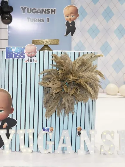 A close-up of the fluted cake table for a Boss Baby theme. The table is decorated with pampas grass and a character cutout, with the baby's name in lights at the base.