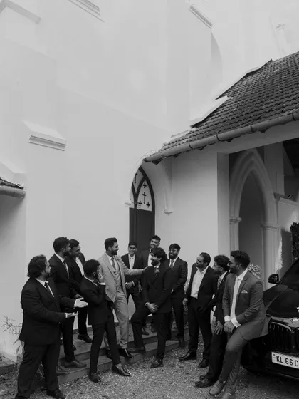 The groom shares a laugh with his groomsmen outside the church. This candid black and white shot captures the relaxed and happy atmosphere among friends before the ceremony.