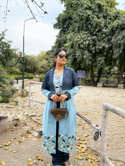 Holding a classic brown leather purse that grounds the blue tones of the denim and floral jacket, showing how accessories can tie a fusion look together.