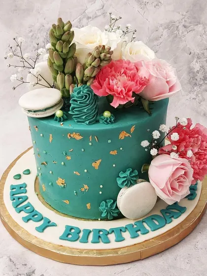 We combine the beauty of succulents and flowers in this unique green floral birthday cake. A rich teal cake is the perfect backdrop for a lush arrangement of fresh pink and white flowers, edible succulents, and macarons, creating a vibrant, botanical look.
