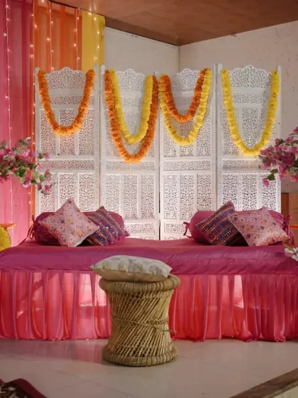 An indoor Mehendi seating area with a beautiful white carved screen backdrop. The space is decorated with marigold garlands and plush pink seating for a traditional yet comfortable feel.