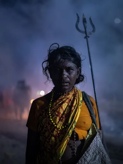 A woman holding a trishul stands amidst the smoke and haze of a festival. Her powerful silhouette embodies a raw, divine feminine energy.