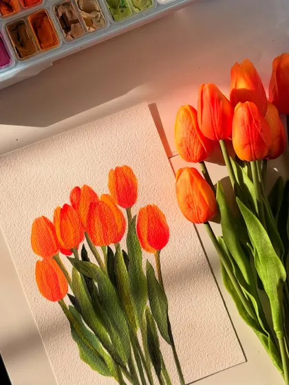 The finished orange tulip painting, resting next to the real flowers and my gouache palette. The warm sunlight in this shot really brings out the vibrancy of the colors.