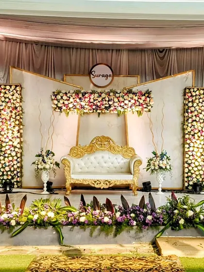 A beautifully symmetric wedding stage design featuring panels of white flowers and a central floral header. The elegant white sofa and personalized nameplate complete this classic and timeless look.