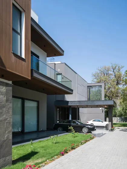 The linear volume of Triveni Farmhouse is broken up by projecting elements and a covered carport, creating a dynamic and functional facade. The mix of dark grey stone, white plaster, and warm wood adds texture and visual interest.