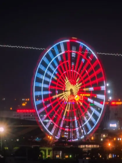 A long-exposure photograph of a brightly lit Ferris wheel in Vietnam, with the light trail of an airplane flying behind it.