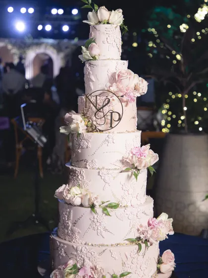 A six-tiered blush pink wedding cake, personalized with the couple's monogram. The design features delicate lace piping and cascading sugar flowers, creating a timeless and romantic look for a grand wedding.