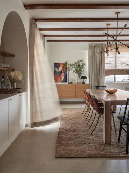 In this dining room, we used a long wooden table and exposed ceiling beams to create an earthy, rustic feel. The sheer curtain, caught in the breeze, adds a sense of movement and lightness to the space.