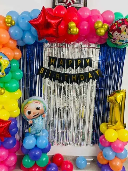 A colorful Cocomelon themed first birthday party. This home setup includes a balloon arch over a foil curtain, character cutouts, and a number "1" foil balloon.