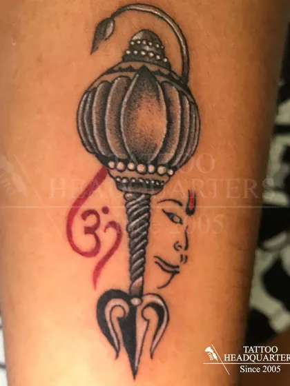 A composite spiritual tattoo featuring Hanuman's Gada (mace), a Trishul, the Om symbol, and a depiction of a deity's face.