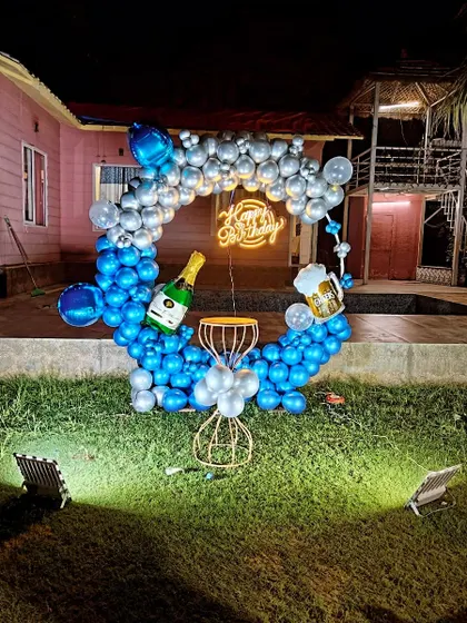 A fun outdoor birthday party setup on a lawn. This features a circular balloon ring in blue and silver, a 'Happy Birthday' neon sign, and playful foil balloons of a champagne bottle and beer mug, ideal for a relaxed celebration.