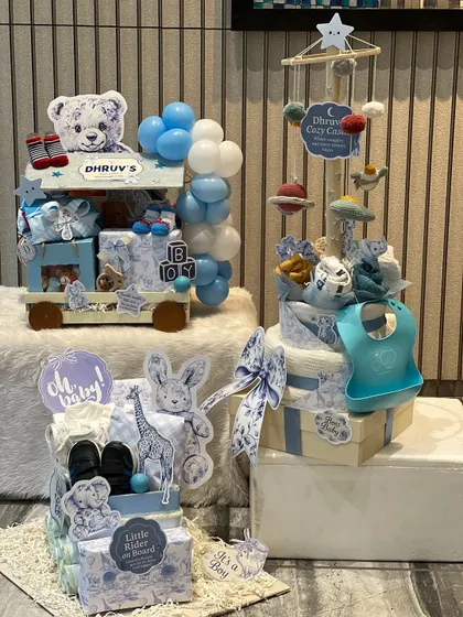 An overview of a custom baby arrival collection for a boy named Dhruv. It includes a trolley hamper, a diaper cake with a cozy mobile, and a mini wardrobe, all in a coordinated blue and white print.