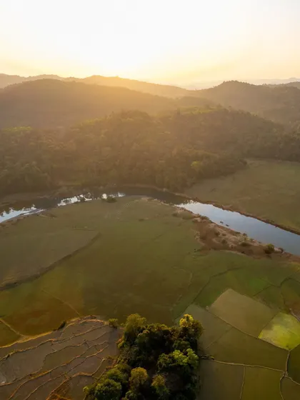 The warm light of sunset over the fields and river in the Western Ghats. This top-down perspective captures the serene beauty of rural Karnataka.