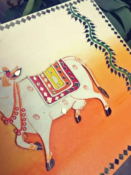 A traditional Lippan art piece featuring a sacred cow against a warm, sunset-colored background. The cow is adorned with mirror work, celebrating its significance in Indian culture.