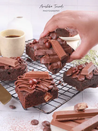 A hand holding a rich KitKat brownie, styled with coffee and more brownies on a cooling rack.