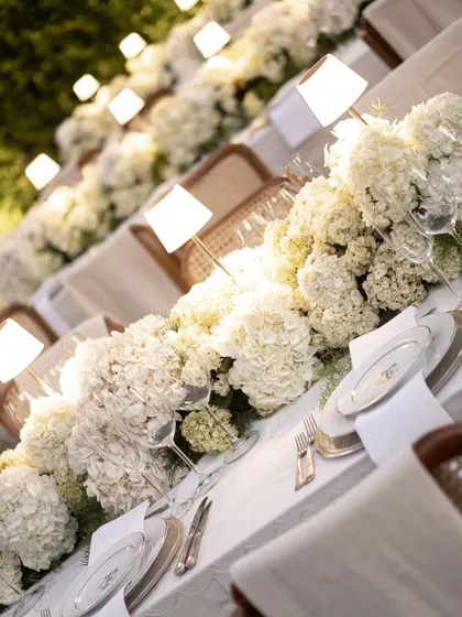 An angled shot of a long dining table, emphasizing the rhythmic placement of glowing lamps and floral arrangements. This creates a visually stunning and cohesive look for a large dinner party.