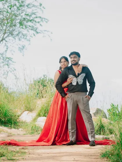A beautiful outdoor shoot. Her red tail gown drapes elegantly, while his modern black shirt with a butterfly motif adds a unique touch.