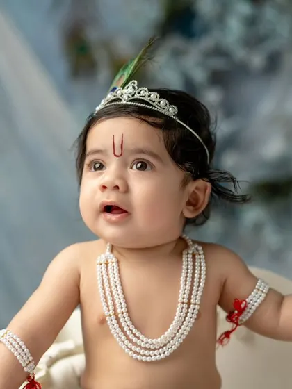 A curious little Krishna looking up in wonder. The soft, dreamy blue background with peacock feathers creates a beautiful and ethereal mood for this baby portrait.