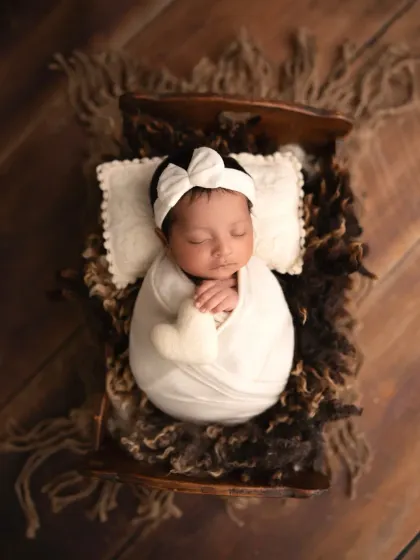 A classic newborn portrait. This baby girl is swaddled in white and sleeping peacefully on a tiny wooden bed with a heart-shaped pillow.
