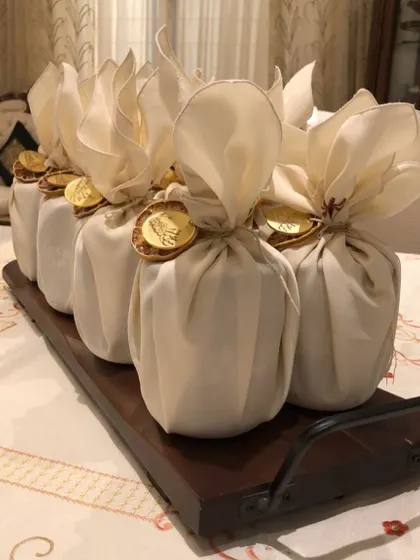Cheesecake jars wrapped as elegant Eid favors. Each jar is wrapped in cream-colored fabric and tied with a gold coin, presented on a simple wooden tray.