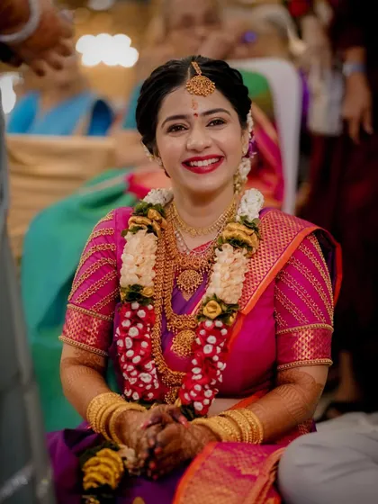The bride, adorned in a stunning purple and pink Kanjeevaram saree and traditional gold jewelry, smiles for the camera.
