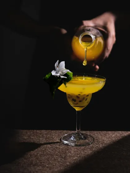 A dramatic action shot of a passion fruit drink being poured, using a single light source to create deep shadows and highlight the splash.