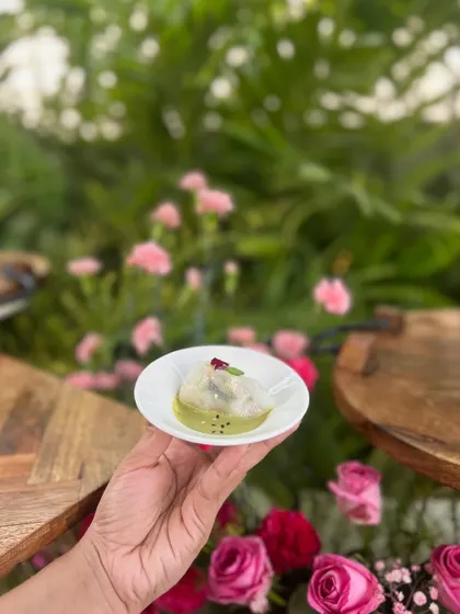 A delicate Thai Basil Dumpling, held in hand against a backdrop of pink roses, from a poolside Haldi celebration.