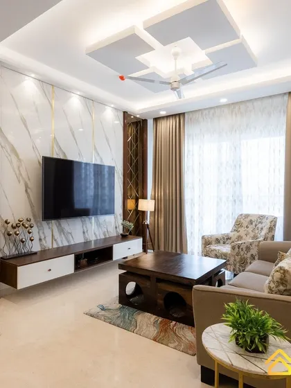 A luxurious living room featuring a floor-to-ceiling marble-finish TV wall with gold inlay details. The design is complemented by a geometric false ceiling and a mix of patterned and solid color seating.