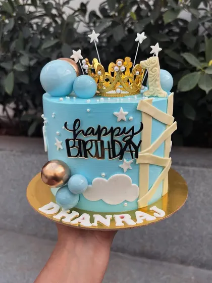 A dreamy blue cake for a first birthday. It's decorated with a crown, stars, clouds, and balloons for a whimsical "prince" theme.