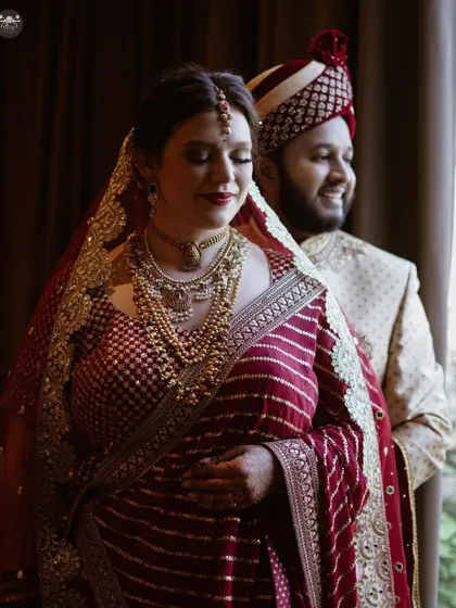 A softly lit portrait of a couple standing by a window. The natural light creates a gentle and romantic mood, highlighting the bride's intricate saree and the couple's serene expressions.
