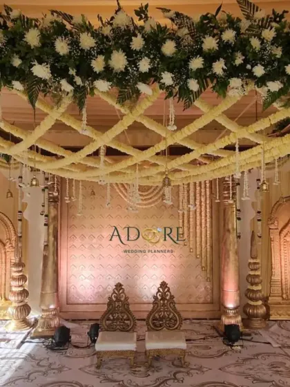 An elegant golden mandap ready for the Muhurtham. The ceiling is decorated with a lattice of white flowers, and ornate statues of deities frame the stage, adding a touch of divine grace.