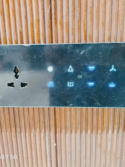 Here is our smart switch panel with a dark, textured finish, blending perfectly with a wooden wall. The blue backlighting makes the icons for lights, fans, and other controls clearly visible.