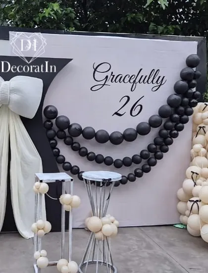 "Gracefully 26." This elegant black and white themed birthday setup is the definition of chic. It features a giant white bow, a string of black pearl-like balloons, and clusters of white balloons with tiny black bow details.