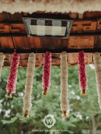 Close-up of the traditional decor elements used in a serene wedding setup. The hanging floral tassels are made of alternating pink and white flowers, ending in small brass bells.
