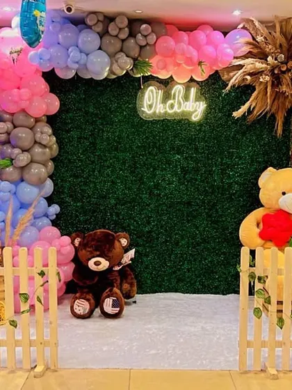 A gender reveal or baby shower setup with a "He or She" theme, using pink and blue balloons, teddy bears, and a green hedge backdrop.