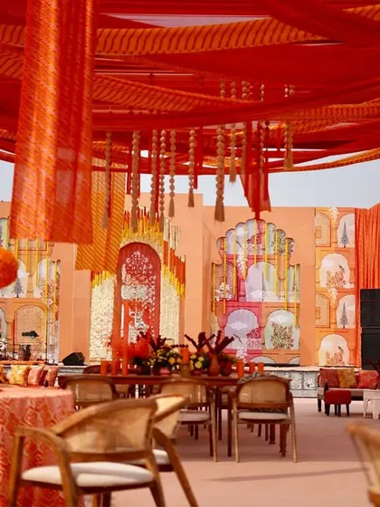 A wide view of the event space, showcasing the hand-painted stage backdrop depicting Rajasthani architecture, the red and orange drape canopy, and the thoughtfully arranged guest seating.