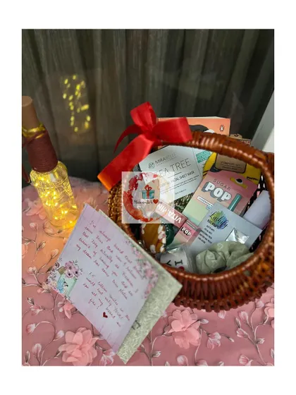 A cozy birthday hamper featuring a handwritten card, a bottle with fairy lights, and a basket full of snacks and self-care items, creating a warm and personal gift.