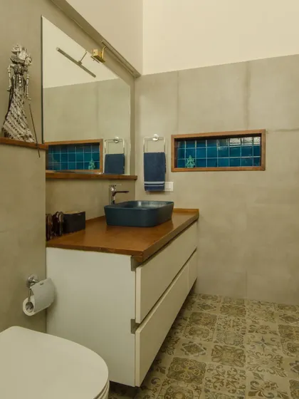 The renovated bathroom combines modern functionality with artisanal touches. A deep blue ceramic sink sits atop a wooden counter, with patterned floor tiles and a niche of bright blue tiles adding color and character.